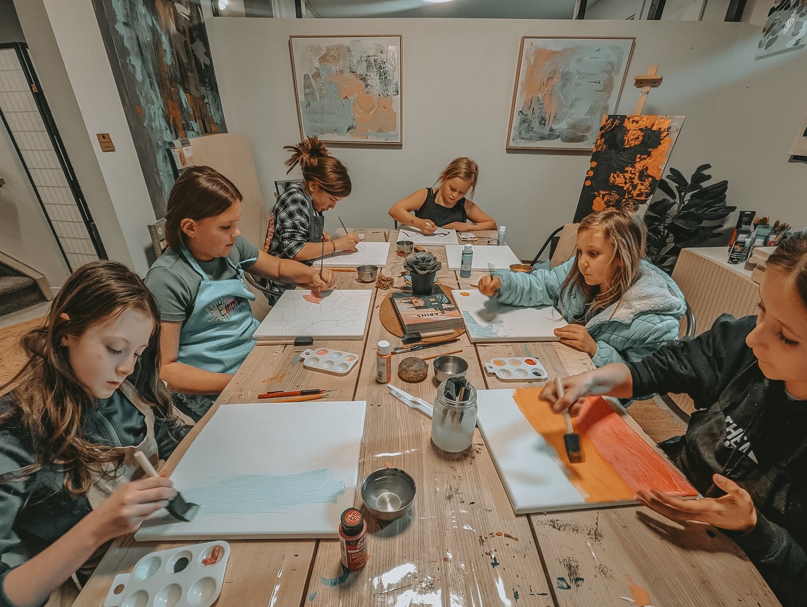 Five girls painting canvases at the long studio table at a Painting Prodigies class, with Leta Taylor's paintings on the walls behind them