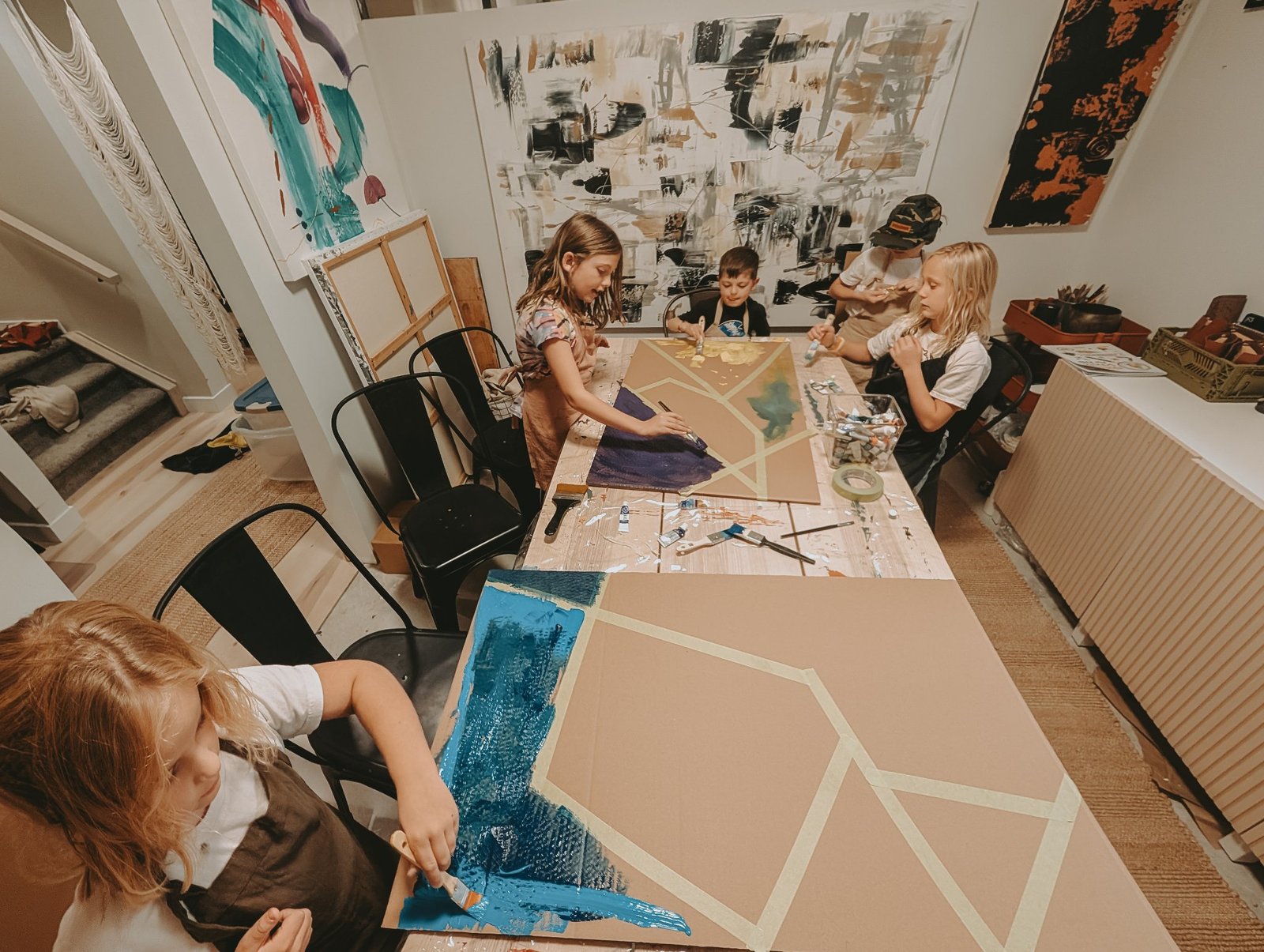 Children painting a large geometric canvas together at the studio table, with Leta Taylor's abstract work on the wall behind them