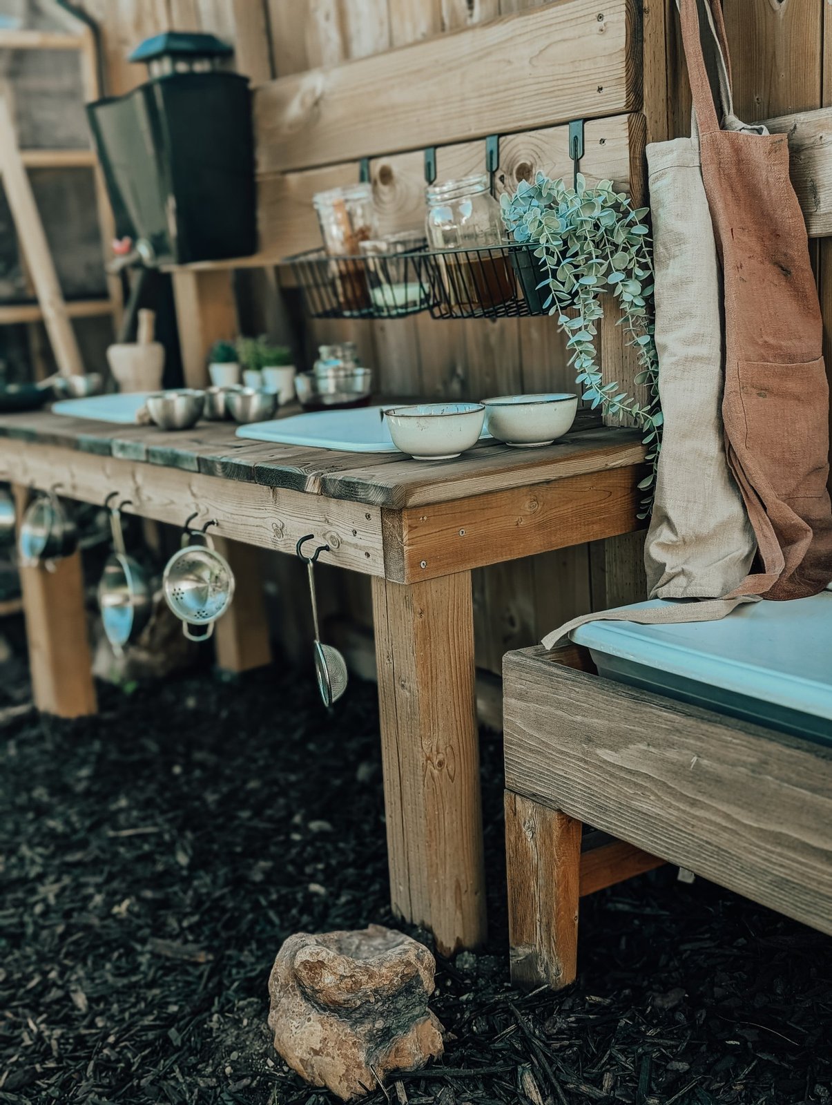 Still life of the outdoor bench: linen aprons hanging beside ceramic bowls and trailing eucalyptus on warm wood, no people