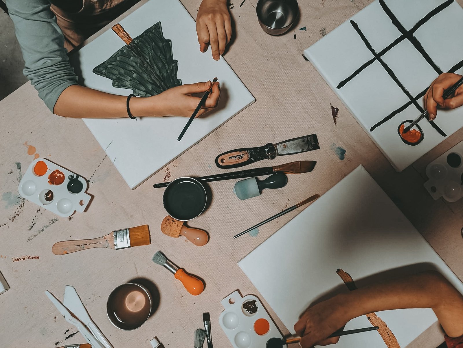 Overhead view of hands painting canvases with palettes, brushes, and palette knives on a linen-covered table, no faces shown
